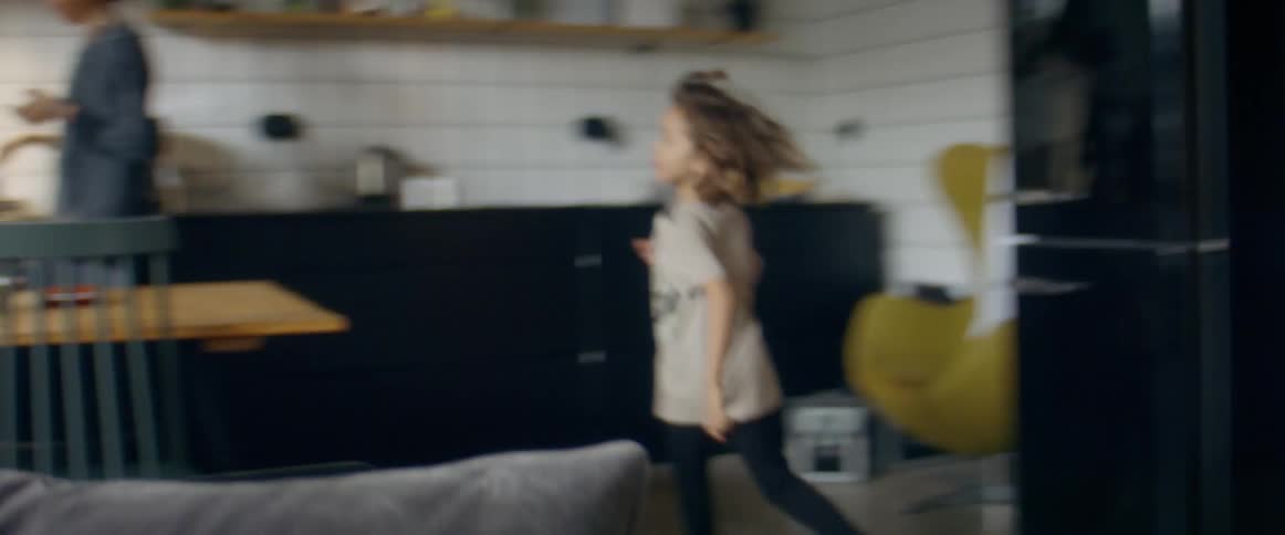 A sister and a brother chasing each other through a living room and a kitchen in a home while their parents watch at daytime