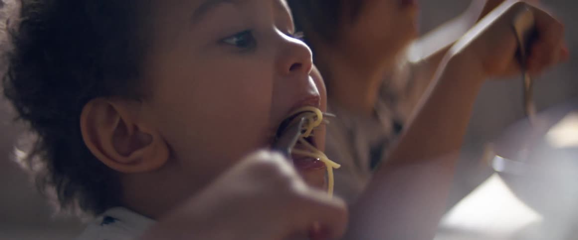 A young boy holds up a fork with pasta and eats next to a sibling