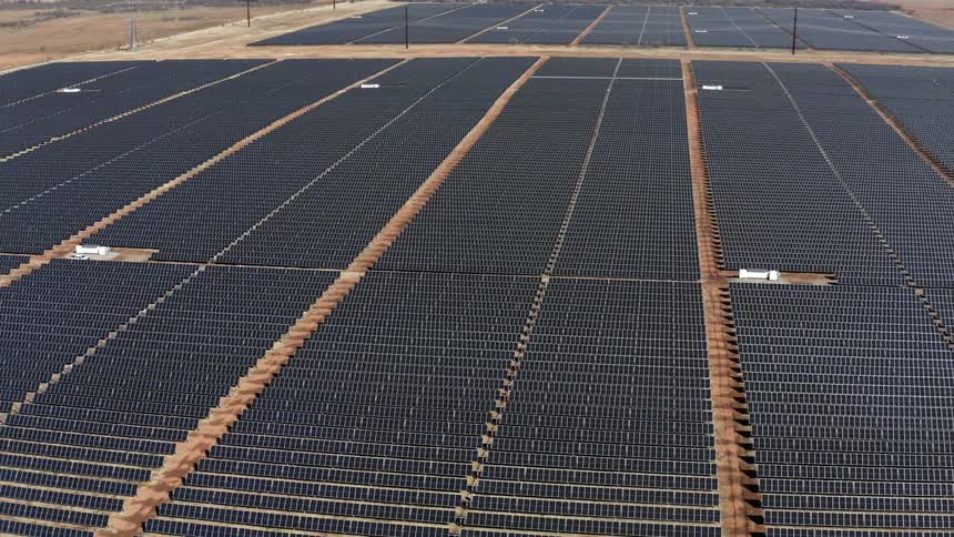 Aerial view of rows of black solar panels reflecting sunlight on a dirt field