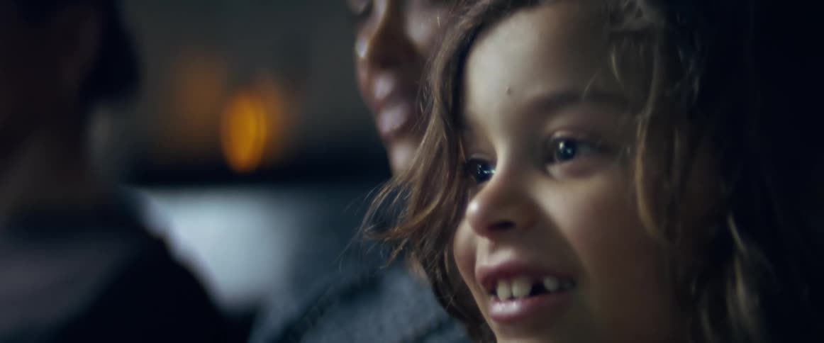 A little boy in a room smiling with two people and a wall in the background
