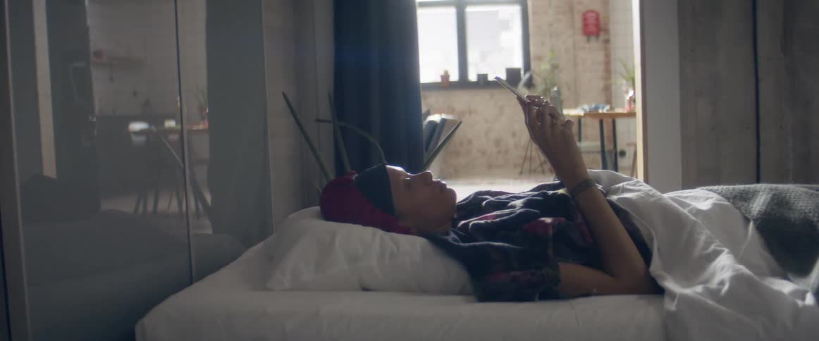 A woman lies in bed at home and looks at her cell phone