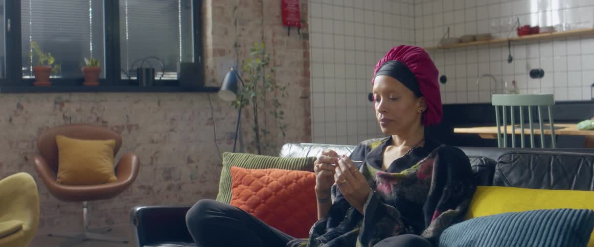 A woman sitting at home on a couch takes a thermometer out of her mouth and looks at it