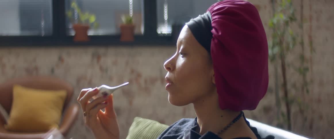 A woman taking her temperature with a mouth thermometer while sitting on a living room sofa