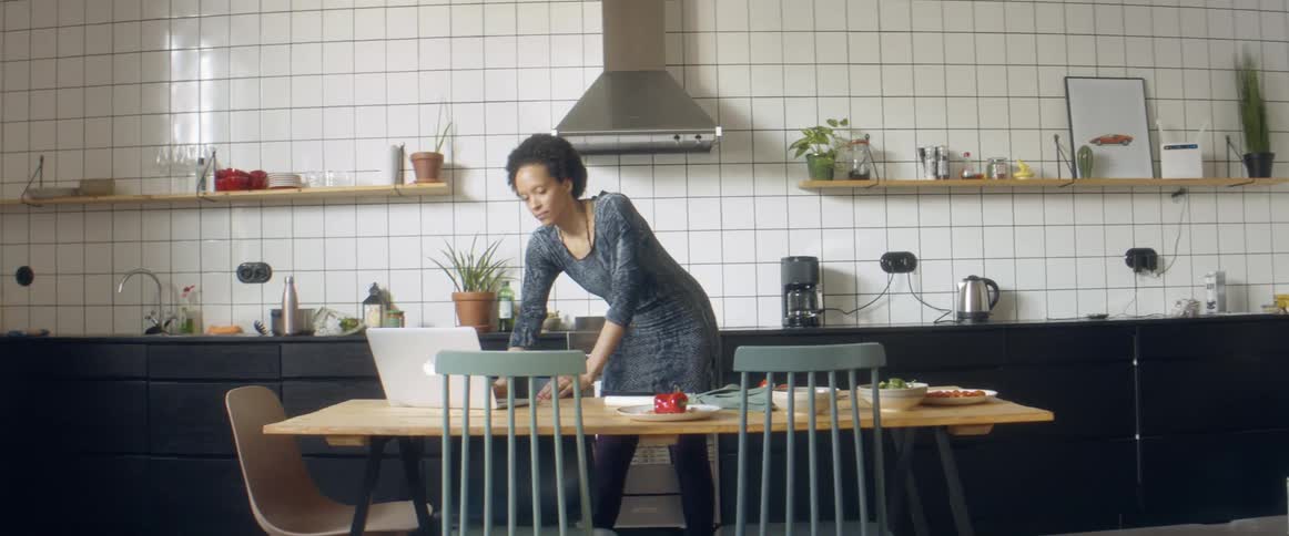Woman with frizzy hair and a blue dress listens to music and cuts vegetables while dancing in the kitchen