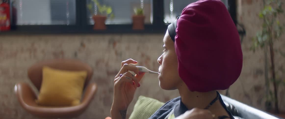 A woman sitting on a couch and putting a thermometer in her mouth with a wall and window in the background