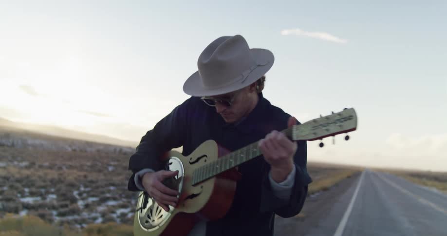 A man walks on the side of a road next to a desert while playing a guitar and singing