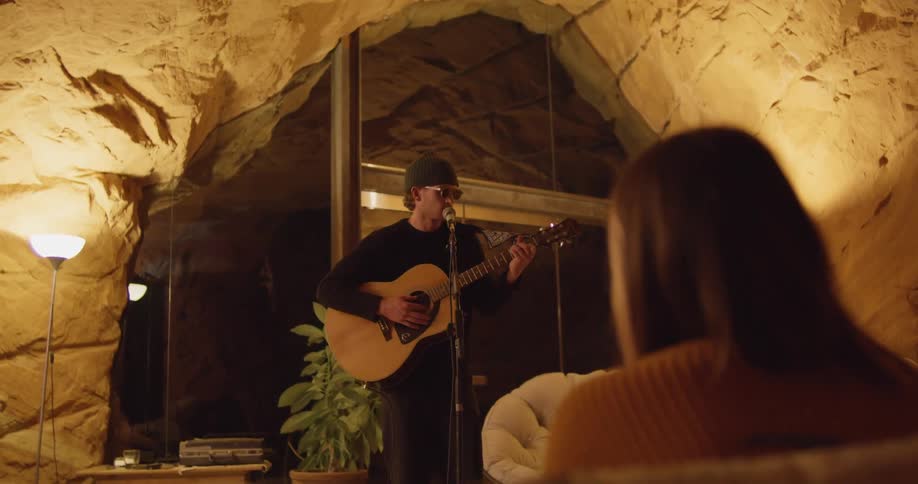 A man standing in a stone archway in a home singing at a mic and playing the guitar while a woman sitting down in front of him watches