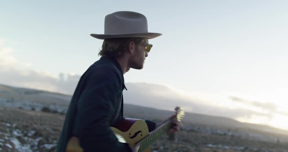 Man with a hat sunglasses and a dark blue shirt plays the guitar in the field at sunset