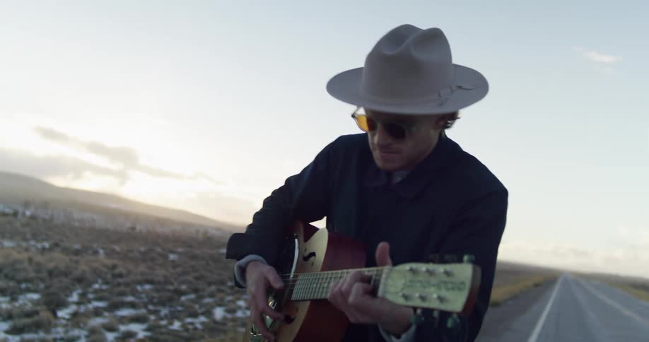 Man playing a resonator guitar while walking on a rural road at sunset