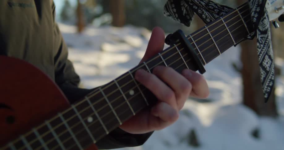 A man with a hat and sunglasses playing a guitar in the forest with snow trees the sky and clouds in the background