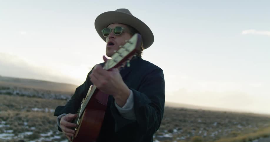A man singing and playing an acoustic guitar while walking alongside a rural road