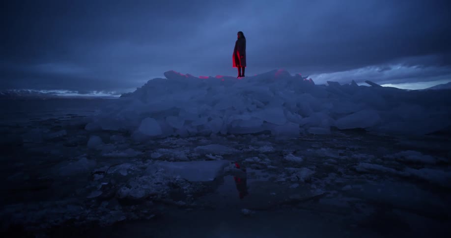 A woman stands on top of a glacier in the ocean as a red light glows below her