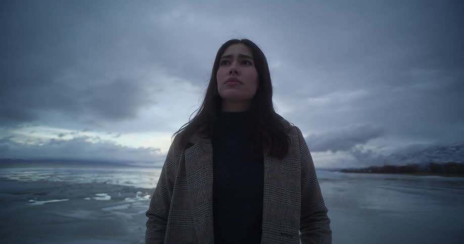 An indigenous woman is standing on a shoreline near ice floating in the water