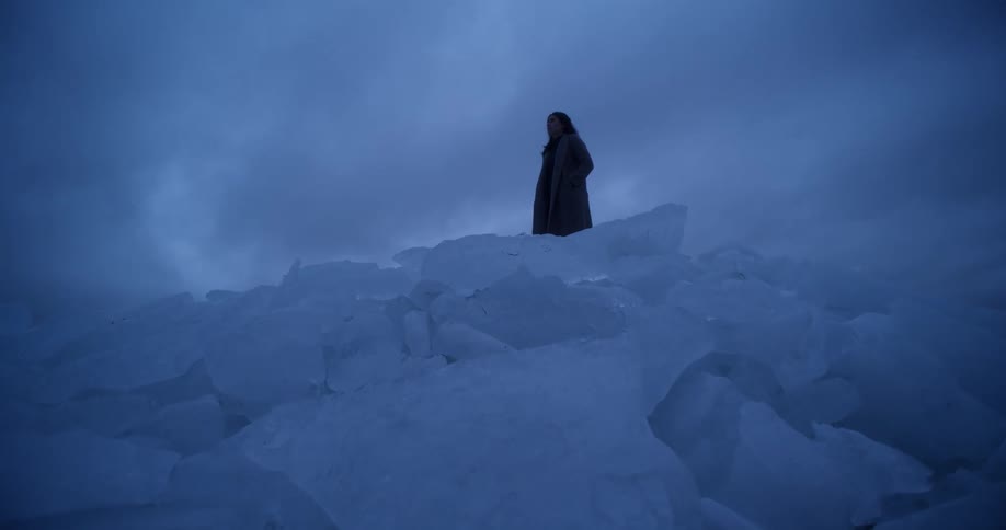 Woman stands at top of mound of large ice chunks under a cloudy sky