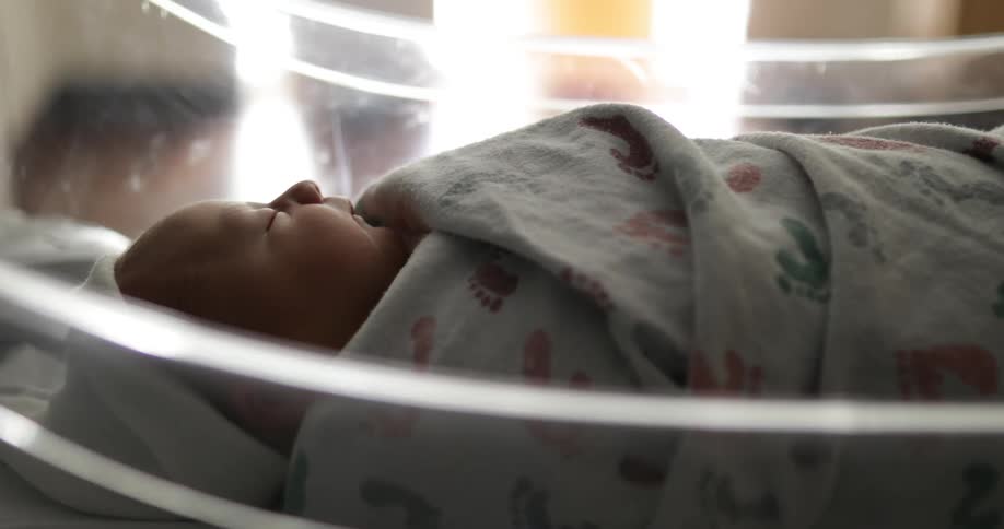 Swaddled baby wearing white cap sleeps in translucent bassinet