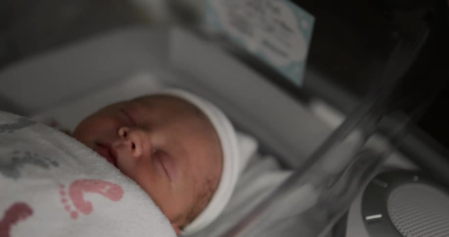 A baby with a hat and blanket laying in a plastic cradle