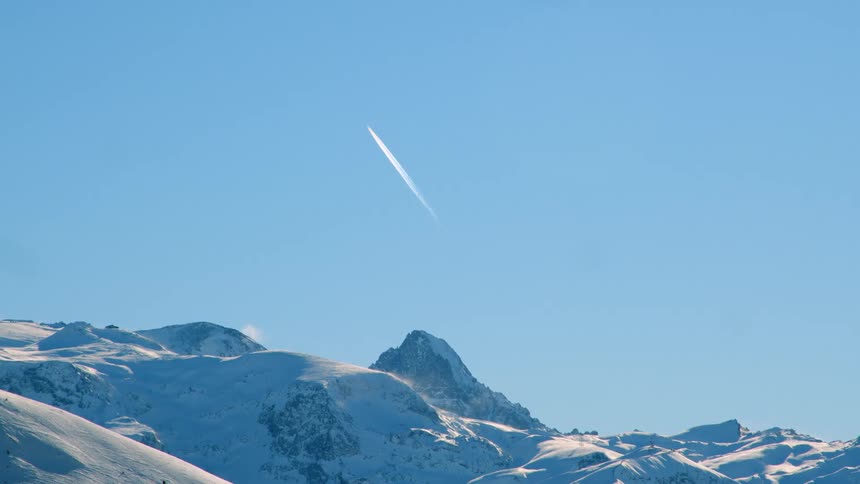 A single contrail blemishes the clear blue sky over a snow covered mountain range