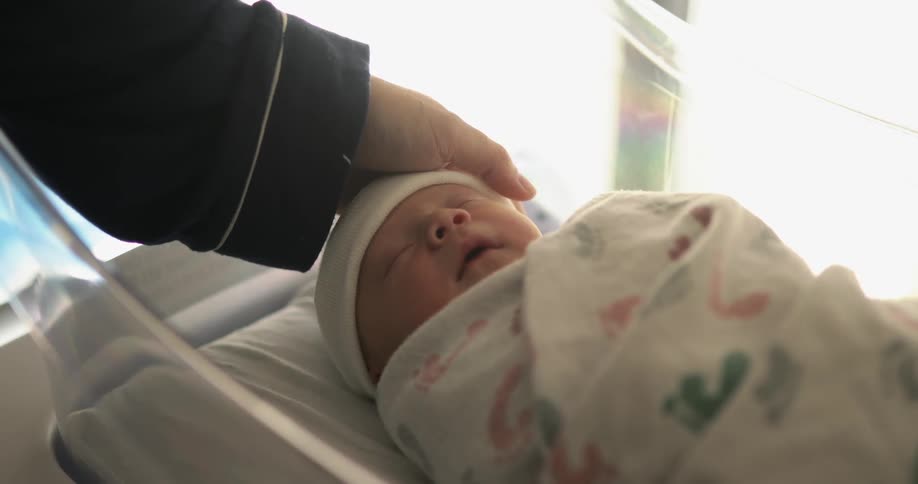 A person caressing a baby with the baby laying in a plastic cradle with a hat and blanket