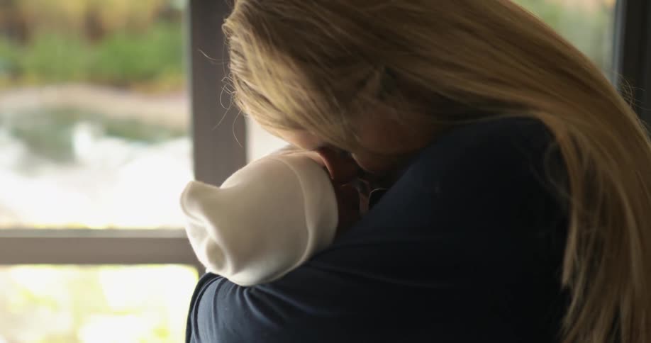 A woman holding and kissing a baby with a hat with a window in the background