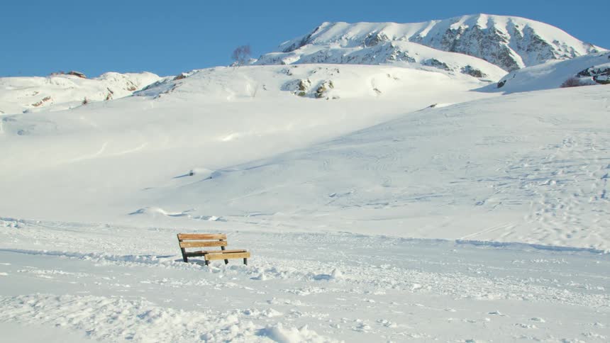 A landscape covered in snow with a bench with a mountain range the sky and clouds in the background
