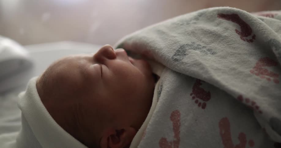 A newborn baby sleeps while swaddled in a blanket with a footprint pattern