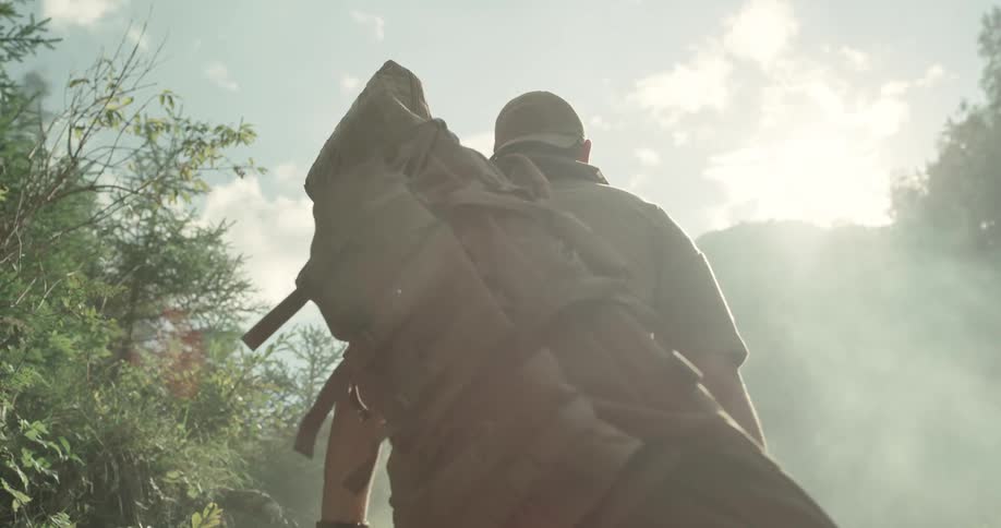 Man carries large pack hiking through forest toward mountain on foggy day under slightly cloudy sky
