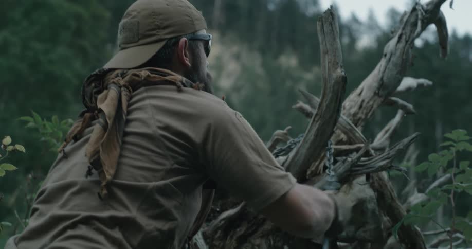 Bearded man hangs object on dead tree with chain with forest topped cliff in background