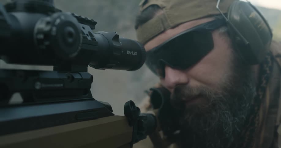 A man with a hat and sunglasses aiming a rifle with a scope