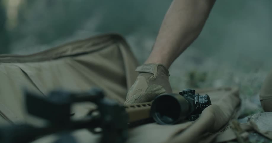 A man in military fatigues kneels on the ground and removes a long rifle from a zipper case