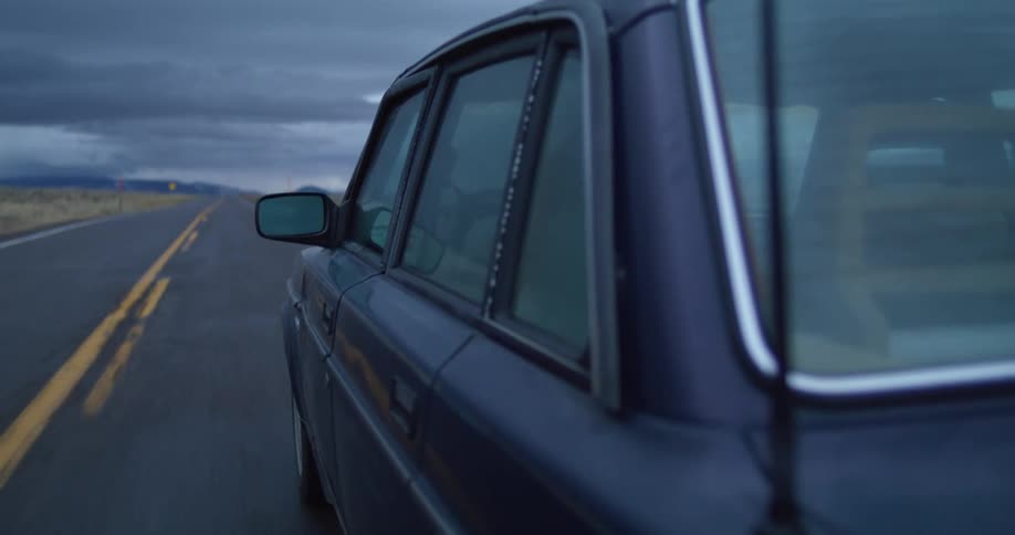 An older blue vehicle drives down a rural road on a cloudy day