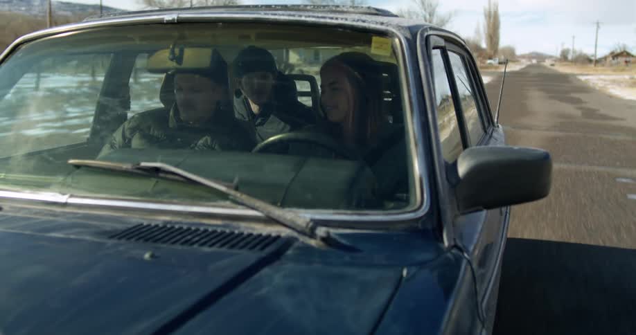 A woman and two men driving in a vehicle on the road with trees the sky and clouds in the background