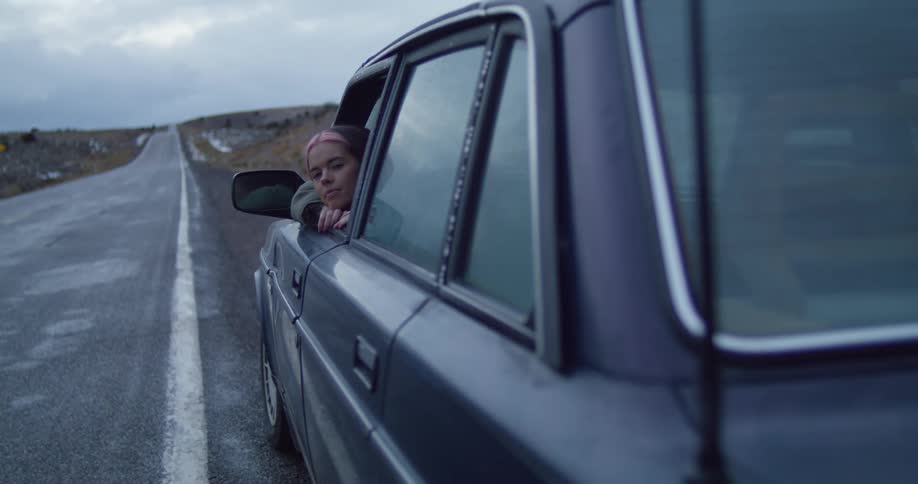 A woman sits with her head out the window of her car as its parked on the shoulder of a rural highway