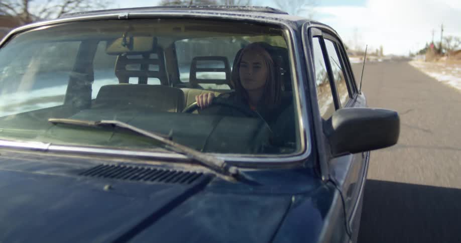 A woman drives an older blue sedan past barren trees on a rural road
