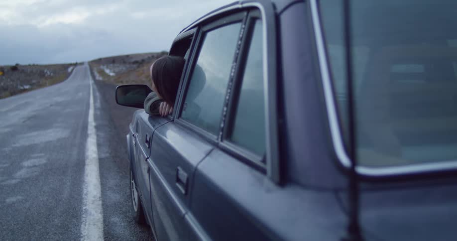 Woman leans out window of car packed on shoulder of road in country looking around under overcast sky