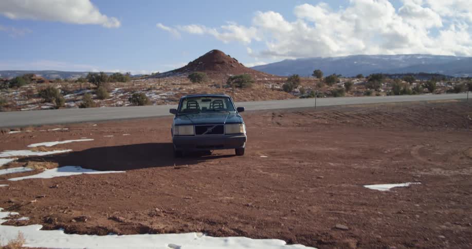 Car sits parked on flat dirt area partly covered with snow across road from mound backed by mountains