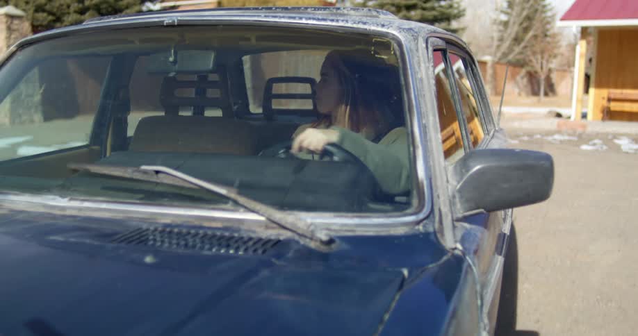 A woman looks to her right to check for traffic as she leaves a parking lot in an older blue vehicle