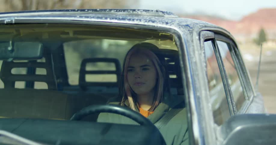 Woman drives along road through country past houses with red mountains in background