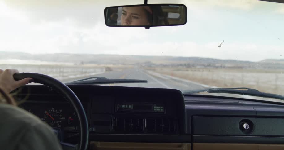 A woman drives down a rural highway with mountains in the distance and melting snow on the ground