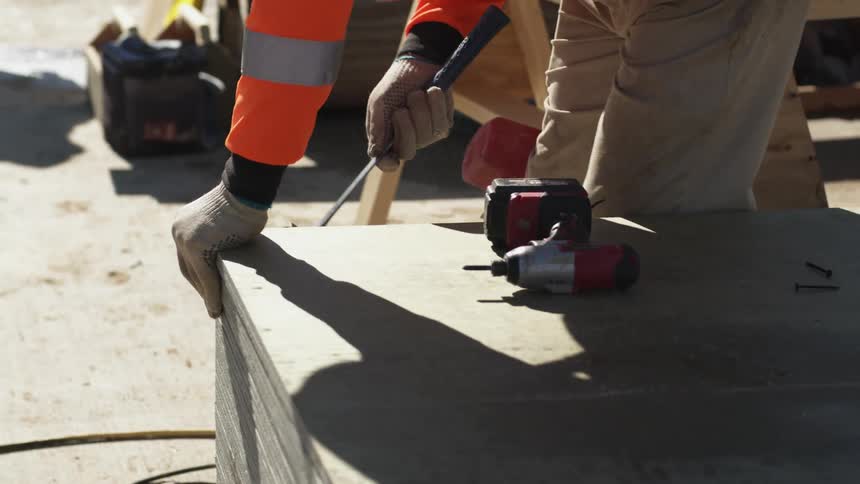 A person with gloves using a hammer on sheet rock with an electric drill on top