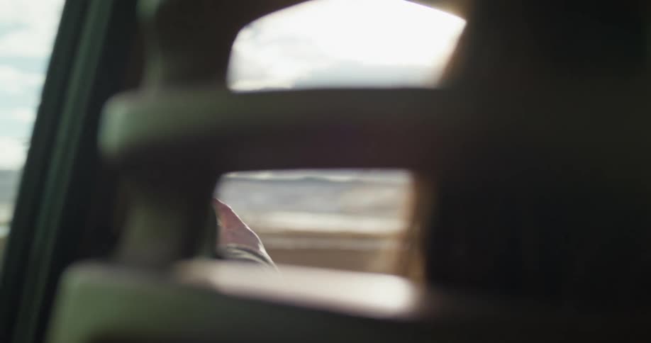 A woman drives down a highway along the ocean with her hand out the of window