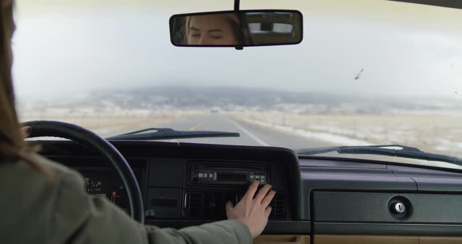A woman driving a vehicle and turning the steering wheel with a landscape the sky and clouds in the background