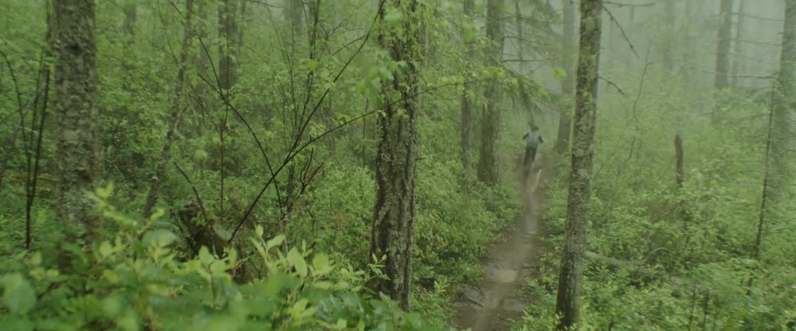 A person riding a mountain bike on a muddy forest trail through puddles and going over a jump