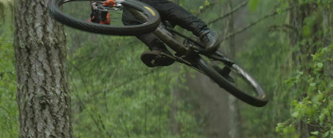 Man wearing helmet races bicycle along dirt trail through forest and whips bike sideways during jump
