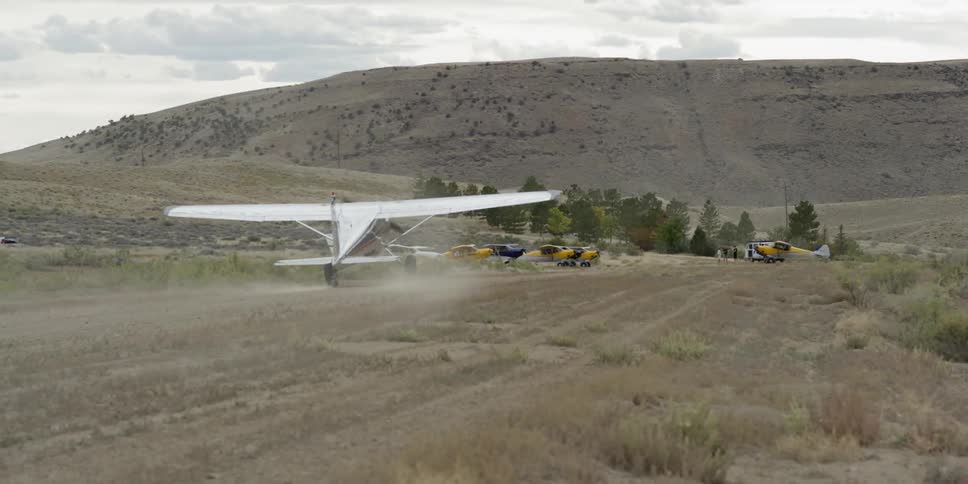 A small plane lands on a dirt runway near a mountain
