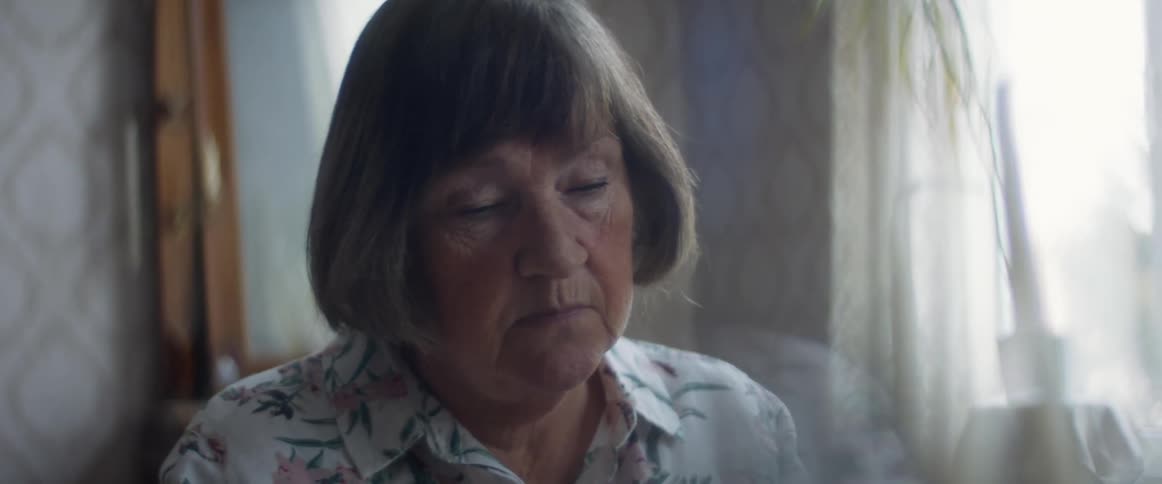 An elderly woman sitting alone indoors takes off her glasses as she concentrates