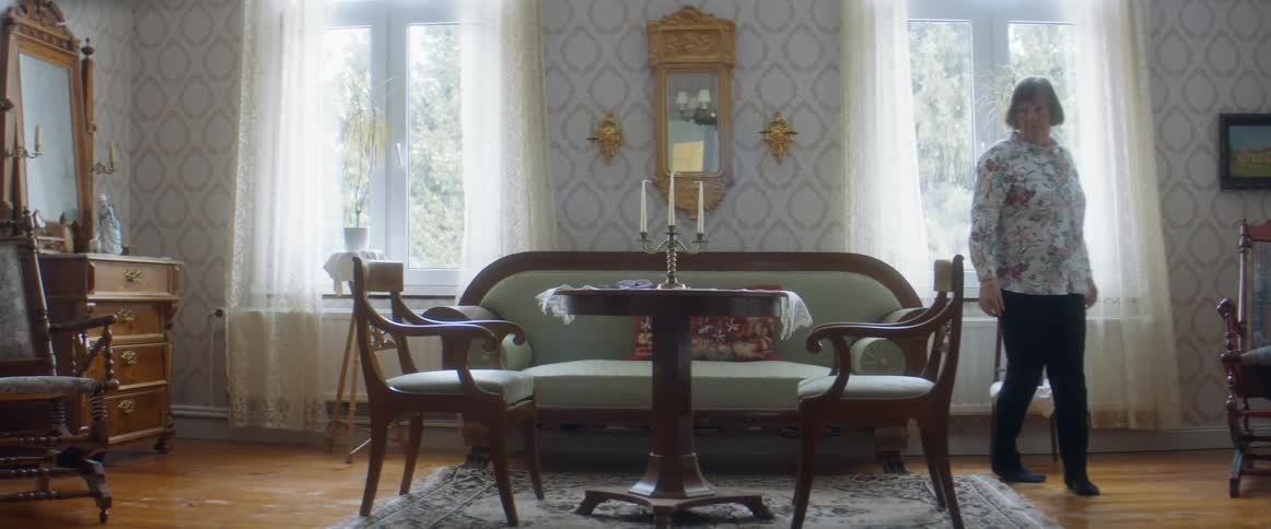 A woman walking in a room with a table and chairs with a wall and windows in the background