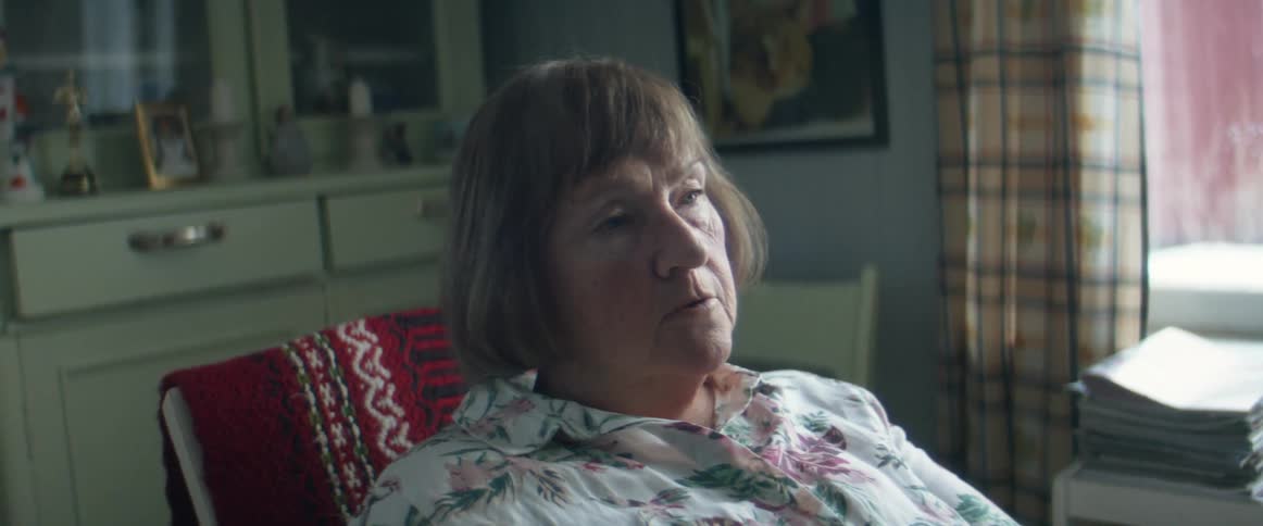 An elderly woman sits in a chair looking depressed and takes off her glasses