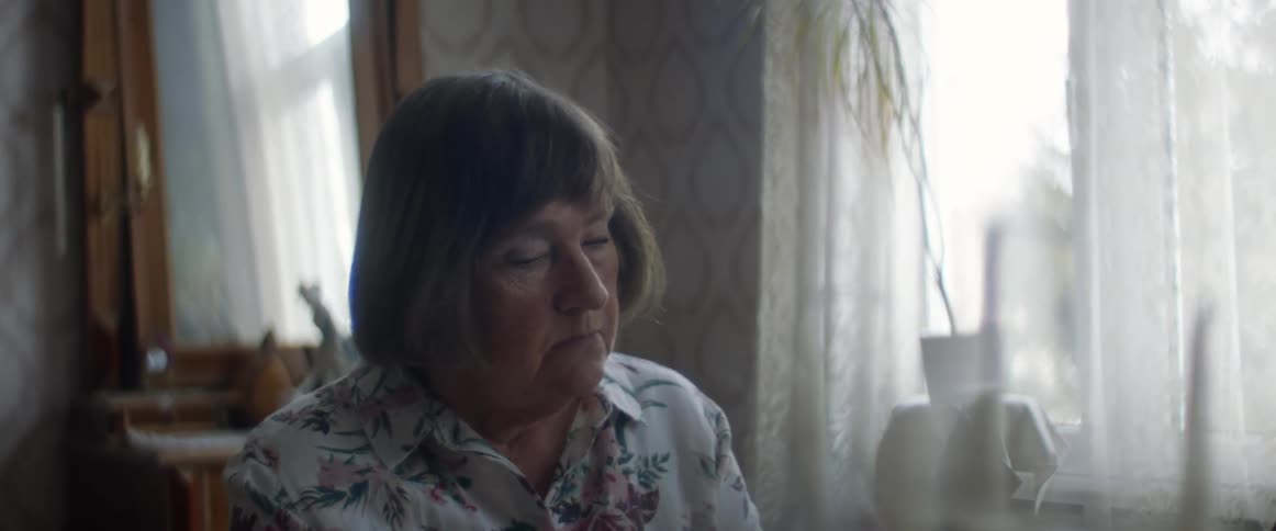 A woman sitting indoors in a bedroom looks up and stares off