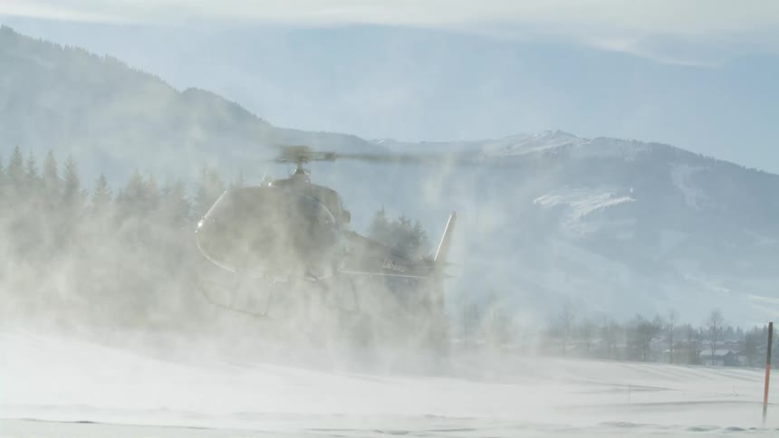 A black helicopter kicks up snow as it takes off from a landing spot near mountains