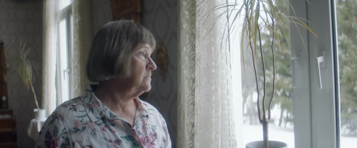 An elderly woman gazes contemplatively out of a window before turning and walking away